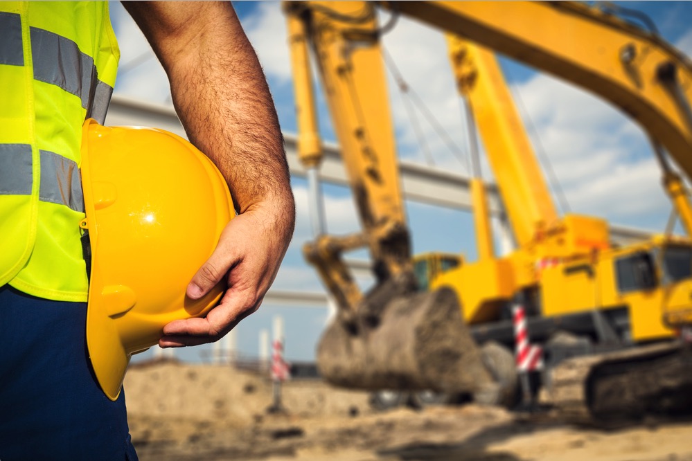 Close up of a man holding a hard hat with an excavator in the background